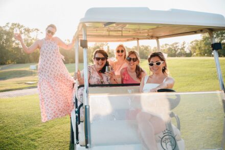 La novia conduce un carrito de golf lleno de amigas con gafas de sol mientras otra posa de pie en el lateral, copa en alto