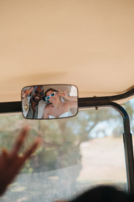 Los novios, con gafas de sol, hacen el signo de la paz reflejados en el retrovisor de un carrito de golf