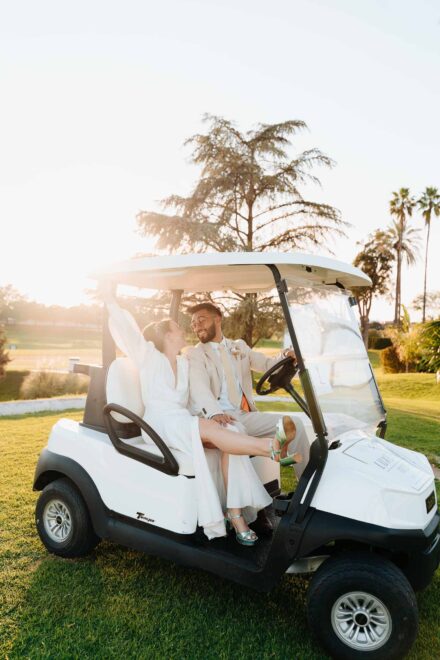 Los novios en un carrito de golf blanco al atardecer; la novia, con sandalias verdes, cruza una pierna sobre él