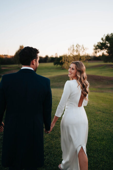 La novia, con vestido de espalda abierta, mira atrás sonriendo mientras camina de la mano del novio por el césped