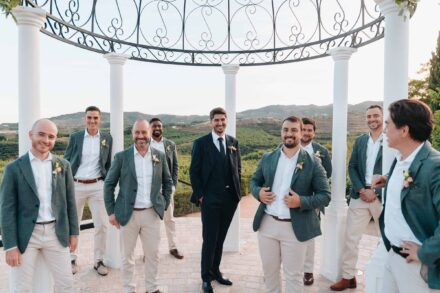 El novio, de traje azul marino, sonríe entre un grupo de hombres con americana verde bajo un templete de forja