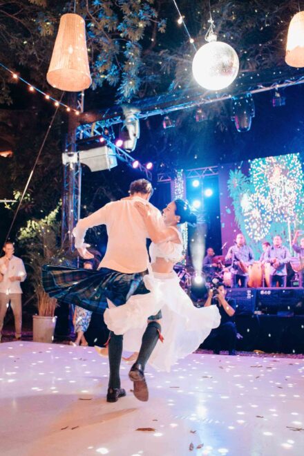 El novio, con kilt, hace girar a la novia en una pista blanca iluminada bajo una bola de espejos, con la banda al fondo