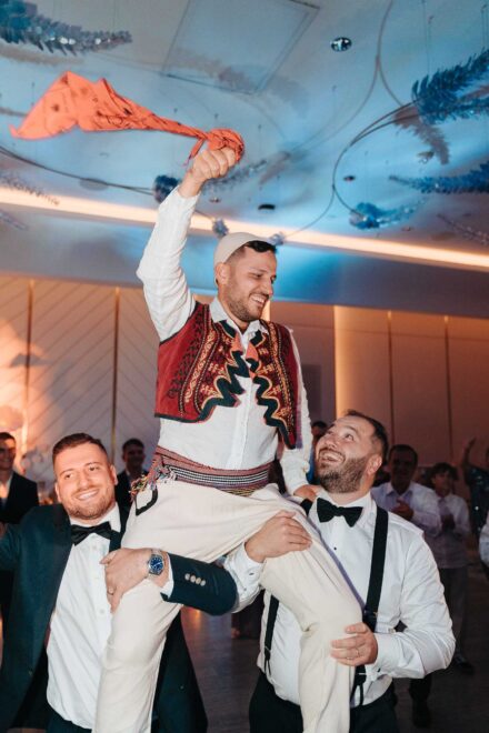 El novio, con chaleco rojo bordado y gorro blanco, agita un pañuelo a hombros de dos invitados con pajarita