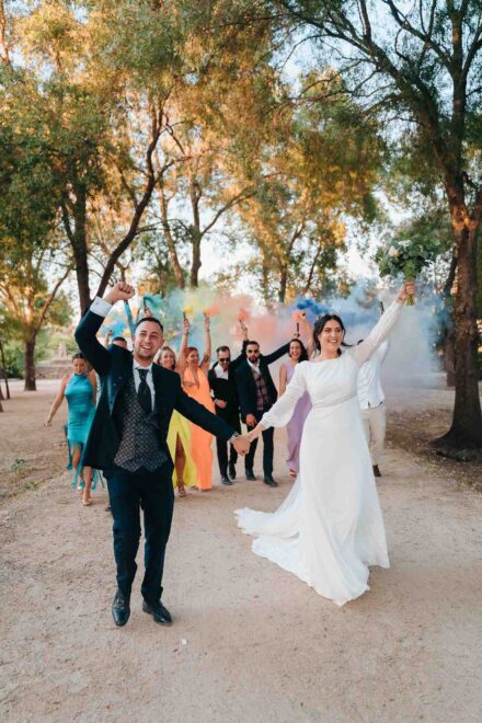 Los novios avanzan de la mano con los brazos en alto, delante de invitados con botes de humo de colores