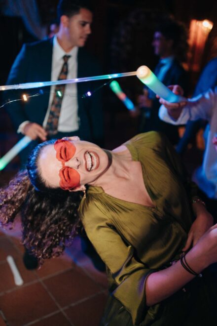 Una invitada con gafas de corazón naranjas ríe echando la melena rizada hacia atrás entre barras de luz