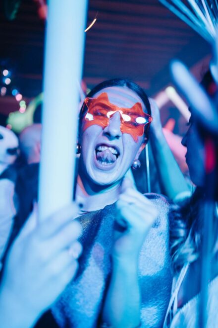 Una invitada con gafas rojas en forma de estrella saca la lengua en la pista, entre luces de neón azules