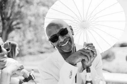 Un invitado con traje claro y gafas de sol sonríe con una sombrilla de papel blanca, en blanco y negro