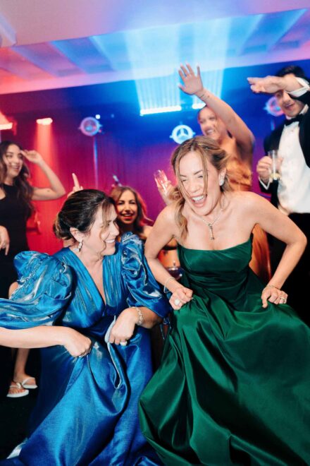 Dos invitadas con vestidos de raso azul y verde bailan agachadas y riendo en la pista