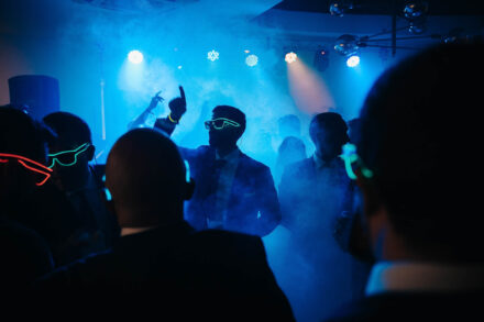 Invitados con gafas de neón bailan a contraluz entre humo azul, uno de ellos con el dedo en alto