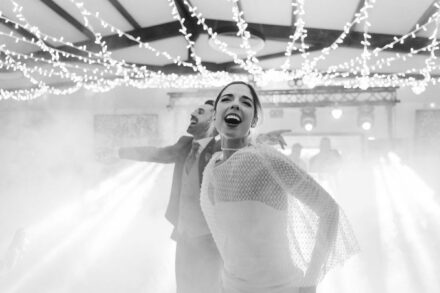 La novia, con capa de rejilla, grita con la boca abierta y el novio abre los brazos detrás, en blanco y negro