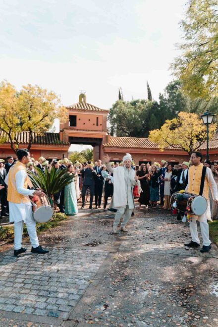 El novio, con turbante y traje blancos, baila entre dos músicos con tambor ante los invitados en un camino empedrado