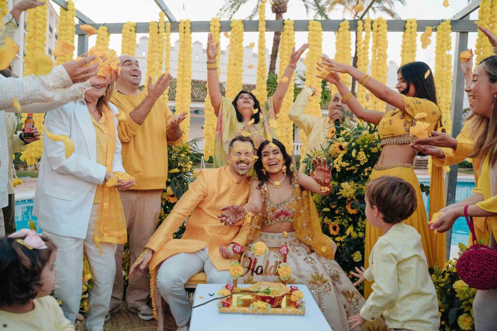 Indian bride and groom laughing during a joyful haldi ceremony, surrounded by family in yellow outfits and flower petals in the air