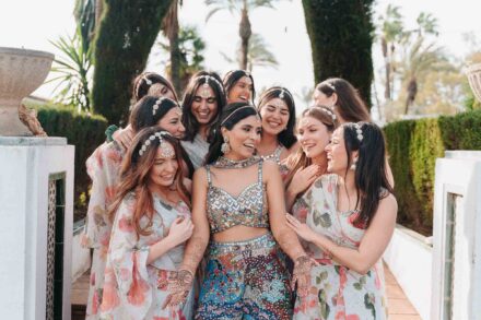 La novia, con conjunto de lentejuelas y henna, ríe rodeada de mujeres con vestidos de flores en un jardín con palmeras