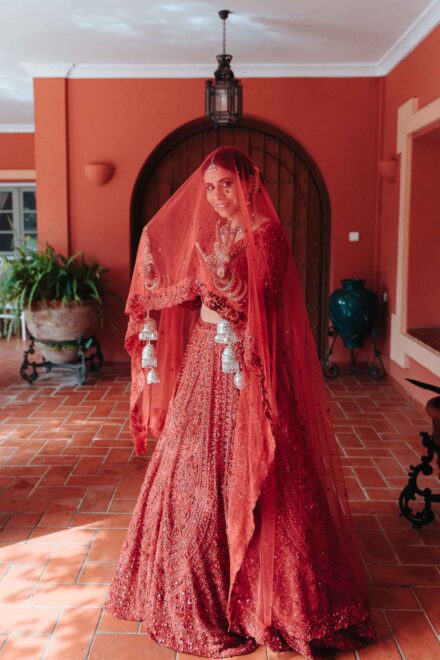 La novia, con lehenga y velo rojos, posa en un patio de suelo de barro frente a una puerta en arco