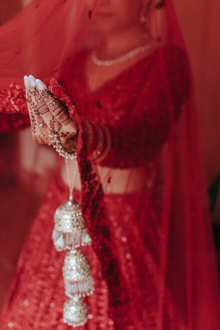La mano de la novia, con henna y kalire dorados, sujeta el borde de su velo rojo de lentejuelas