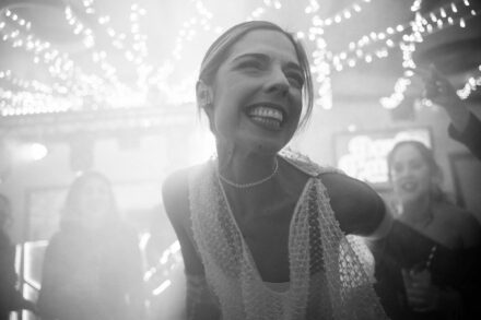La novia, con collar de perlas, ríe a carcajadas mientras baila bajo guirnaldas de luces, en blanco y negro