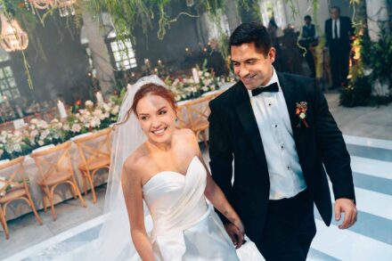 Los novios, sonrientes, caminan de la mano junto a las mesas del banquete, con flores, velas y lámparas de cristal