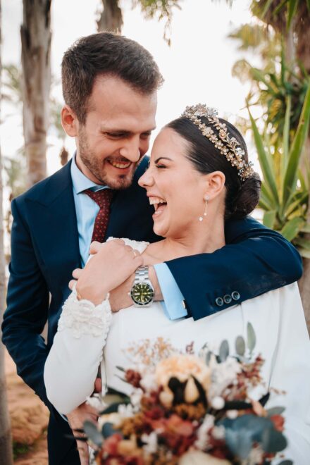 El novio abraza por detrás a la novia, con tiara dorada, mientras ella se ríe a carcajadas con los ojos cerrados