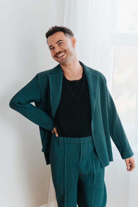 Uno de los novios, con traje verde plisado y camiseta negra, ríe con la mano en la cadera junto a la ventana