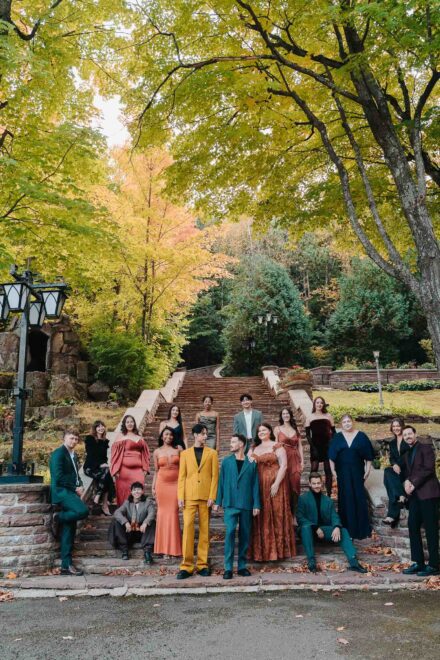 Un grupo con trajes y vestidos de colores intensos posa en una escalinata de piedra entre árboles otoñales