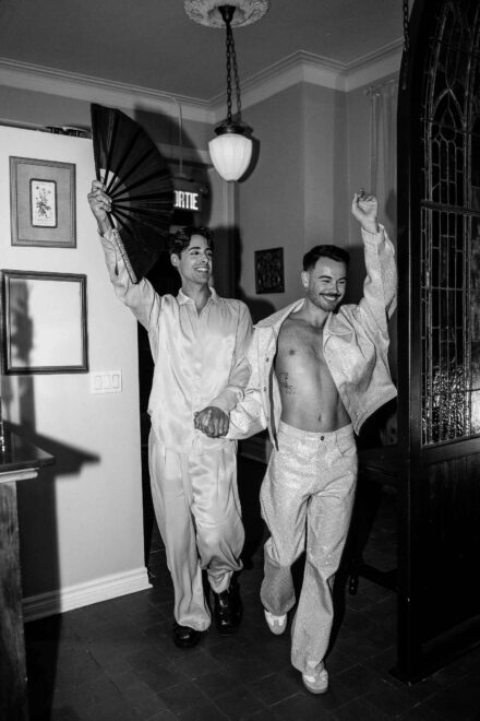 Los novios entran de la mano en la fiesta, uno con abanico negro y el otro con chaqueta de brillos, en blanco y negro