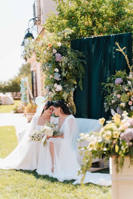 Las novias juntan las frentes en un sofá blanco del jardín, junto a un arco de flores y una cortina verde