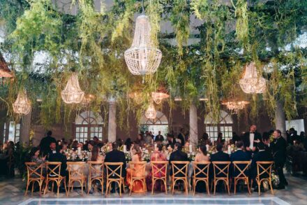 Los invitados cenan en una mesa larga bajo un techo de plantas colgantes y lámparas de cristal