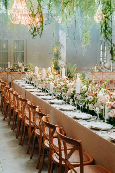 Mesa larga con velas blancas y flores en tonos crema y naranja bajo plantas colgantes, aún vacía