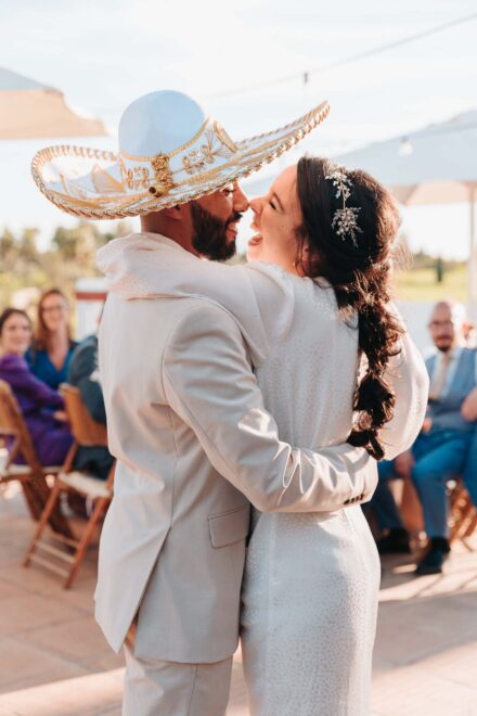 El novio, con un sombrero charro blanco y dorado, abraza a la novia nariz con nariz ante los invitados