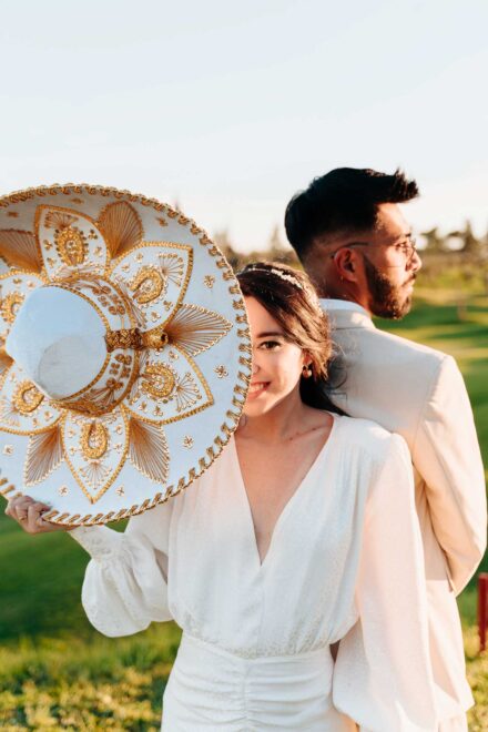 La novia asoma media cara tras un sombrero de charro blanco y dorado, espalda con espalda con el novio