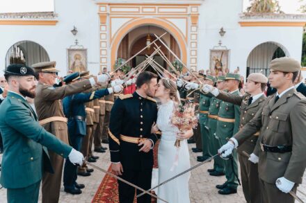 Los novios se besan bajo un arco de sables que forman militares de uniforme, frente a una fachada blanca con arcos