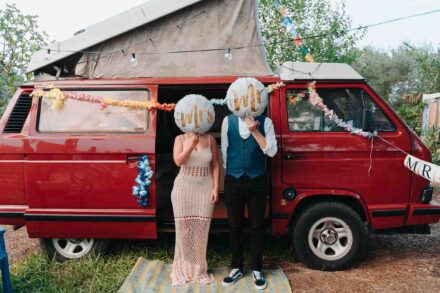 Los novios se tapan la cara con globos de «Mrs» y «Mr» delante de una furgoneta camper roja con guirnaldas