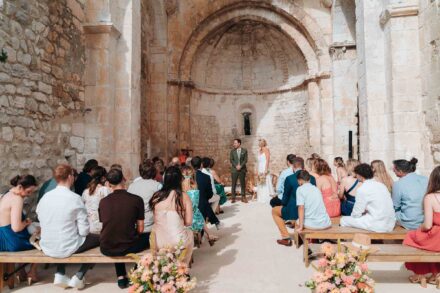 Los novios ante el ábside de una antigua capilla de piedra, con los invitados sentados en bancos de madera