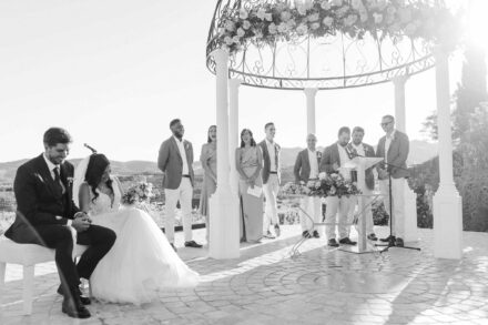 Los novios se ríen sentados en un banco ante un grupo de invitados de pie bajo el cenador, en blanco y negro