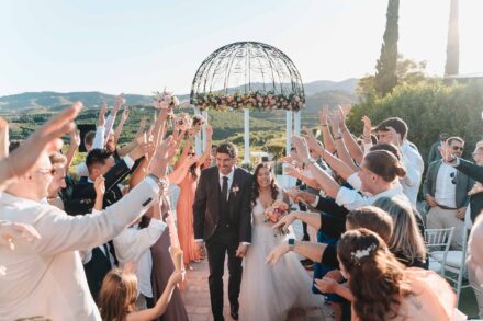 Los novios salen por un pasillo de invitados con los brazos en alto, con un cenador de flores y colinas detrás
