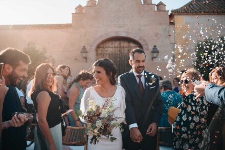 Los novios salen de la mano entre invitados sonrientes y confeti, ante una portada con arco y puerta de madera
