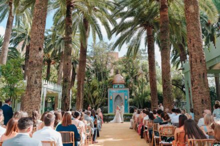 Invitados sentados bajo palmeras altas y los novios al fondo, ante una fuente de azulejos con cúpula