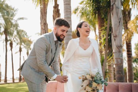 Los novios se ríen de la mano entre palmeras, ella con capa y ramo, él con traje gris de cuadros