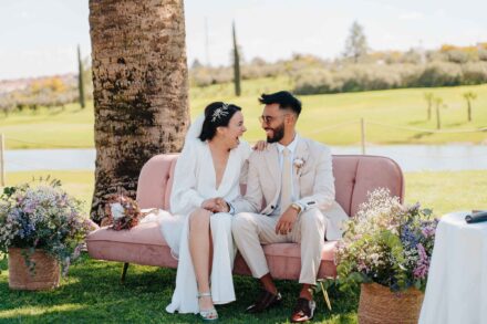 Los novios se ríen sentados en un sofá de terciopelo rosa junto a una palmera, con un lago detrás