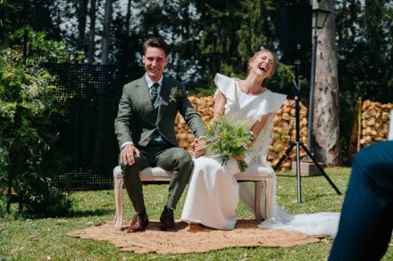 La novia suelta una carcajada con el ramo verde, sentada junto al novio en un banco durante la ceremonia