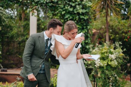 La novia lee sus votos llorando, con micrófono y libreta, mientras el novio le besa el hombro