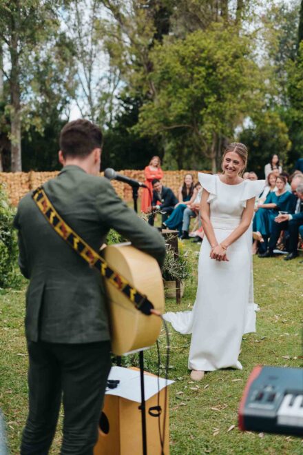 El novio, de espaldas, toca la guitarra ante un micrófono mientras la novia le sonríe en el pasillo