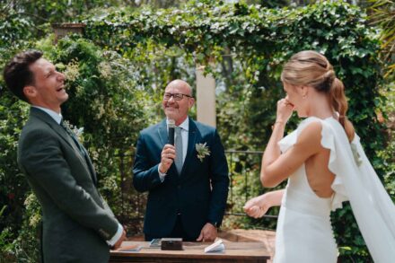 El oficiante habla al micrófono mientras el novio se ríe y la novia se tapa la boca, bajo una pérgola de hiedra