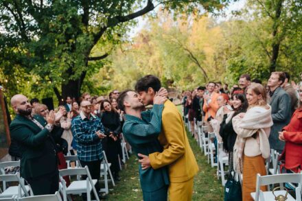 Los novios, uno de traje verde y otro de mostaza, se besan en el pasillo mientras los invitados aplauden