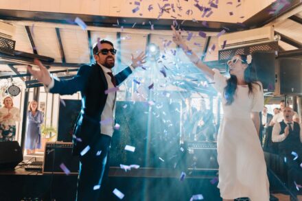 Los novios bailan con gafas de sol bajo una lluvia de confeti morado mientras los invitados aplauden