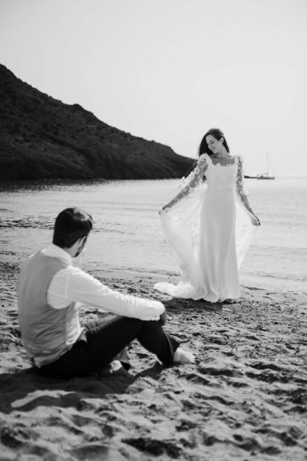La novia abre la capa de tul en la orilla, en la postboda, mientras el novio la mira sentado en la arena