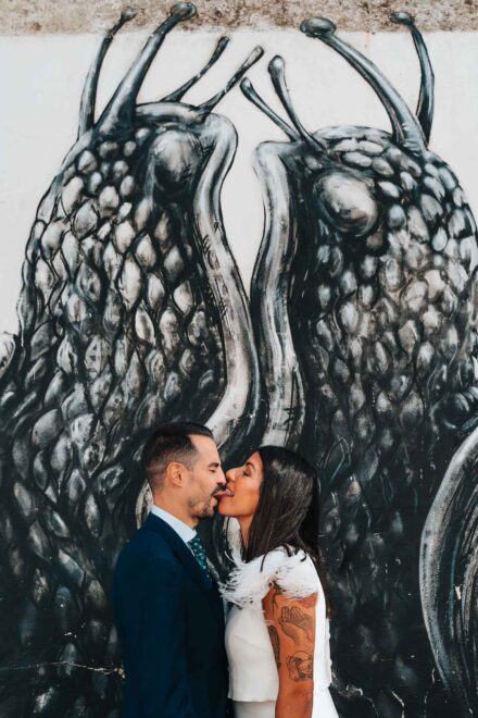 La novia besa al novio sacando la lengua ante un mural en blanco y negro de dos caracoles enormes