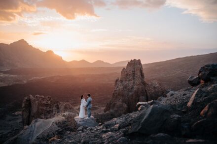 Plano abierto de los novios de la mano en un paisaje volcánico, junto a una aguja de piedra y el sol tras los montes