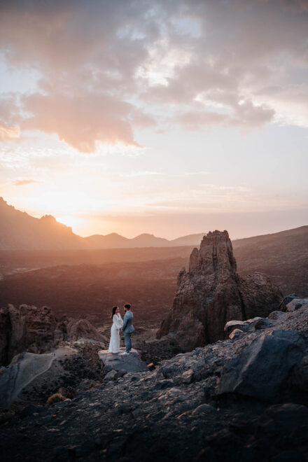 Los novios frente a frente sobre una roca, junto a una aguja volcánica, bajo un cielo de nubes rosadas