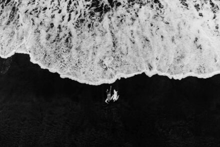 Los novios tumbados en la arena oscura junto a la espuma de una ola, vistos desde el aire, en blanco y negro